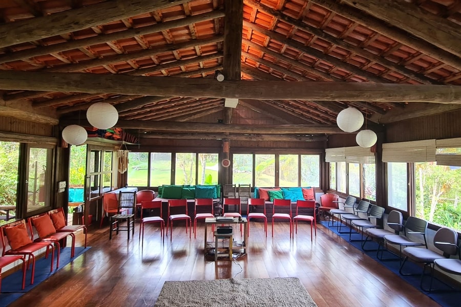 Foto grande sala vazia, cheia de janelas, com piso de madeira e cadeiras vermelhas, organizadas em torno das janelas