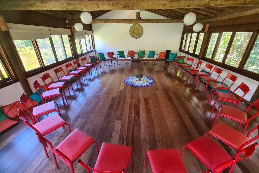 Foto grande sala clara, com piso de madeira, cheia de janelas e com cadeiras vermelhas dispostas em círculo