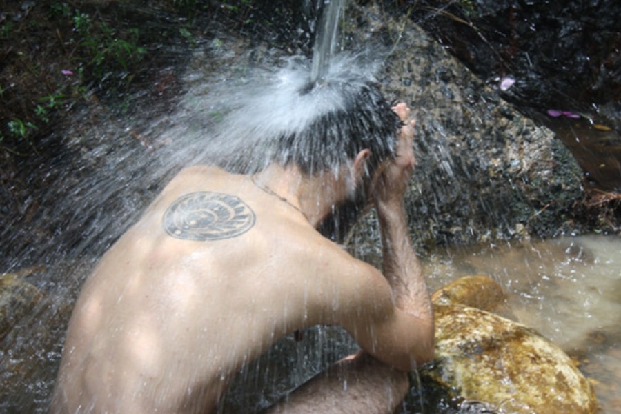 Foto homem agachado de costas na cachoeira, com água caindo na cabeça