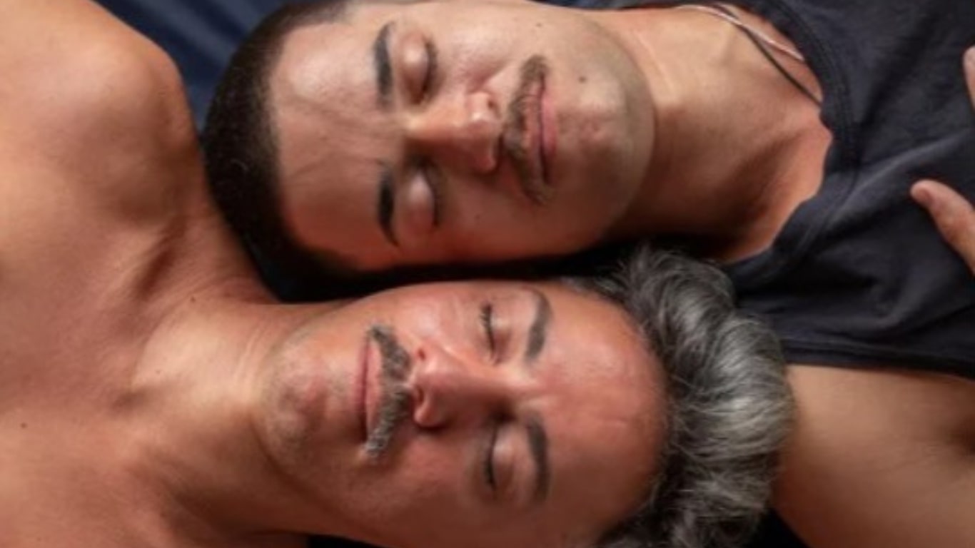 Foto em close de dois homens deitados, com as orelhas encostadas, mas em sentidos opostos, de olhos fechados