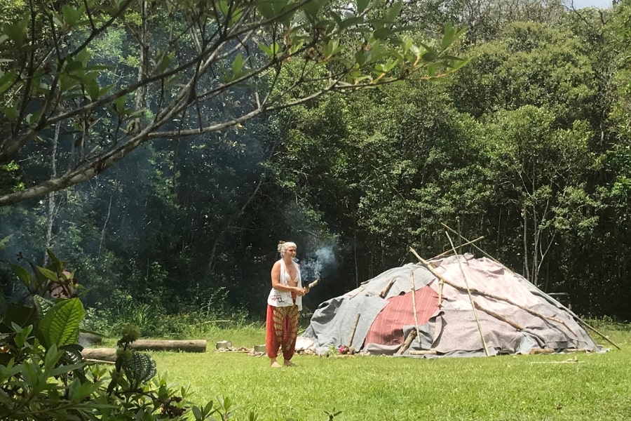 Foto de mulher em pé em frente a uma espécie de cabana, com um graveto aceso