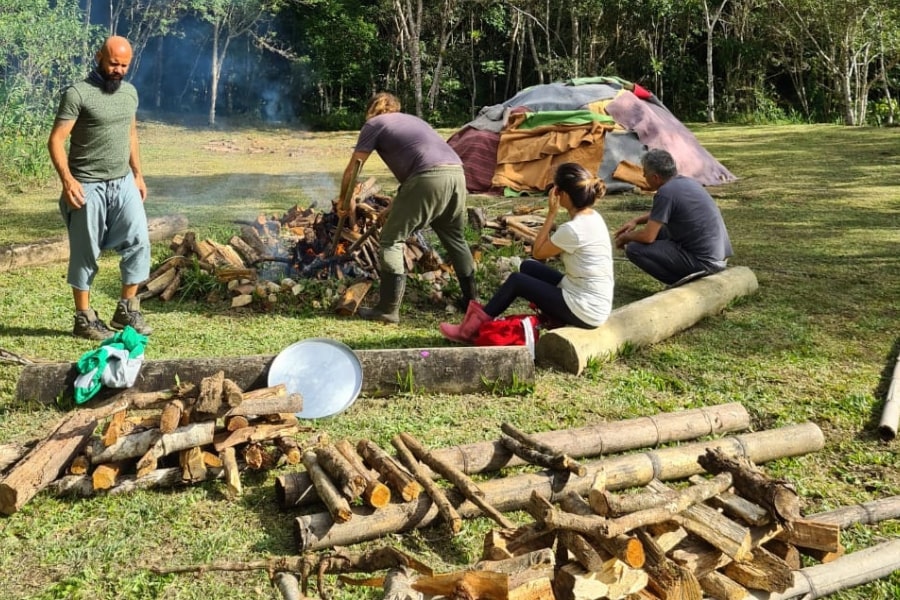 Foto de pessoas trabalhando com lenha para montar uma fogueira, em frente a uma pequena cabana