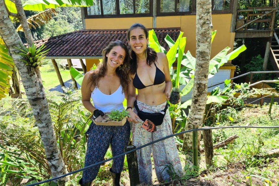 Foto de duas mulheres alegres, posando para a foto, com vegetação e uma casa ao fundo