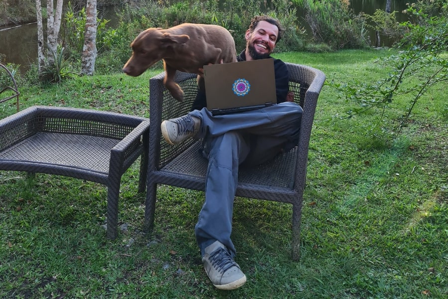 Foto de um homem, sentado em uma cadeira de vime, alegre, com seu notebook e um cachorro grande e marrom