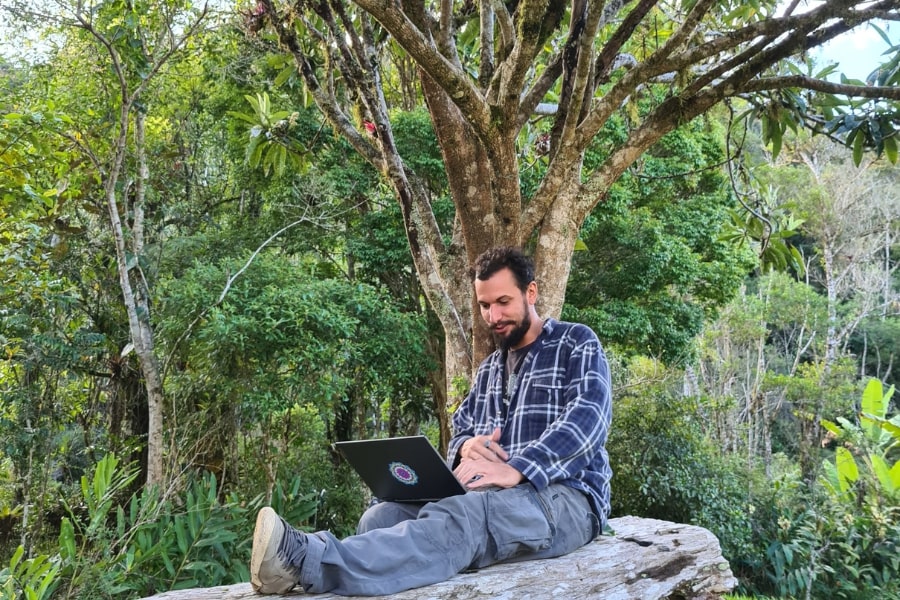 Foto de um homem, mexendo em um notebook, sentado embaixo de uma grande árvore