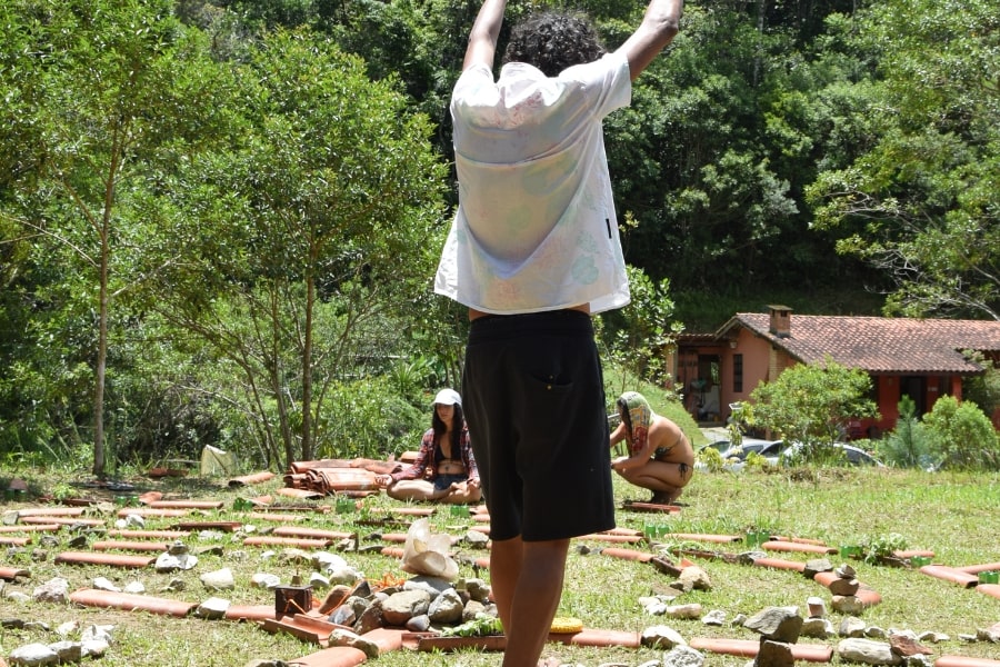 Foto de homem em pé de costas, com os braços para cima, no meio de uma espécie de labirinto de telhas e plantas