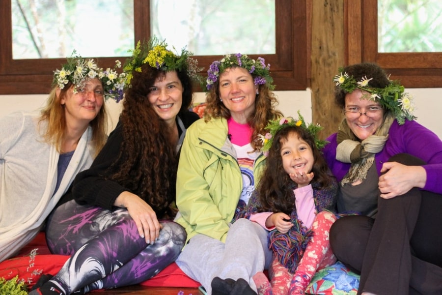 Foto de 5 mulheres sorridentes, com coroas de flores na cabeça