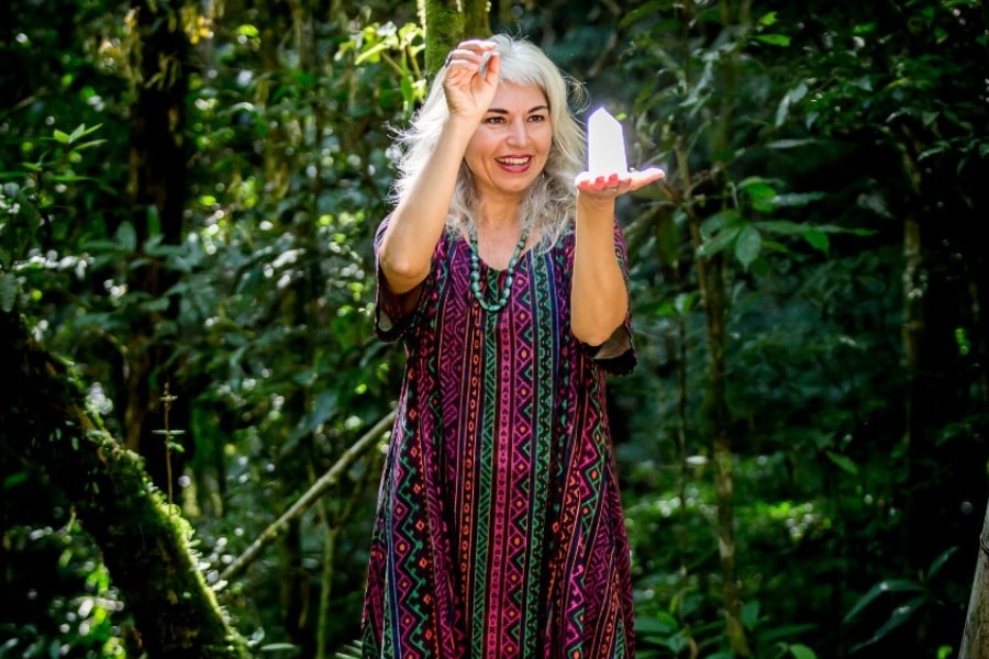 Foto de mulher em pé, segurando um grande cristal na palma aberta de uma das mãos, no meio da floresta.