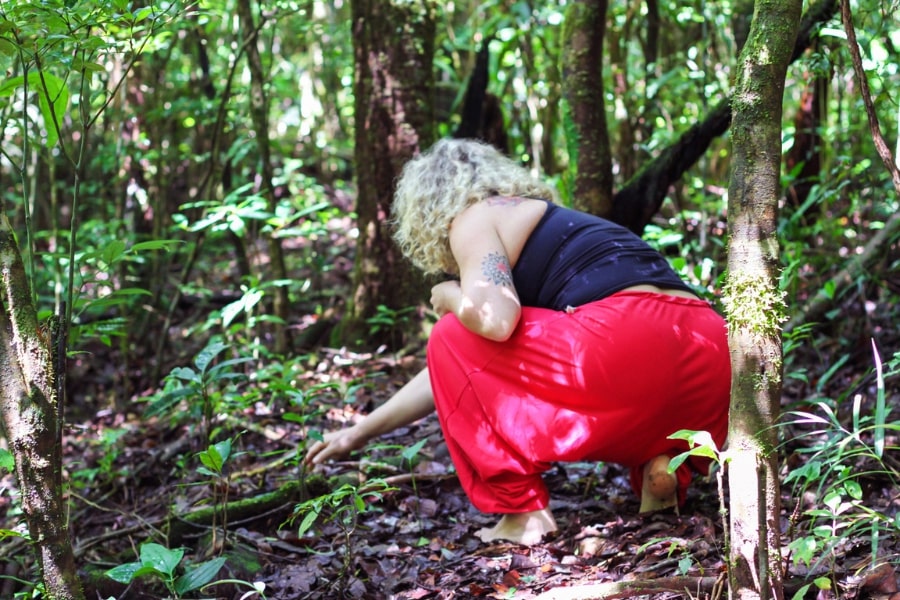 Foto de mulher agaixada de costas, mexendo no solo, na floresta