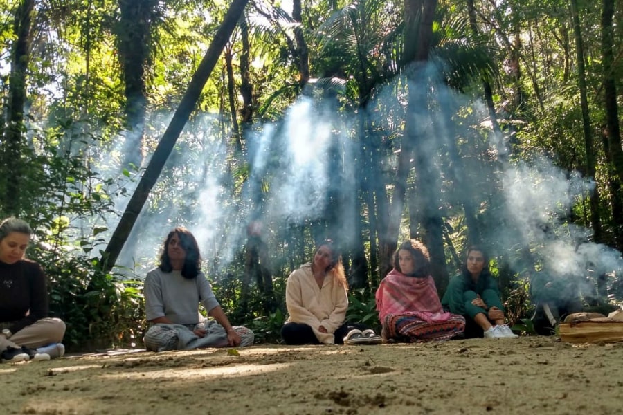 Foto de algumas mulheres sentadas na areia, na floresta, com árvores e raios de sol ao fundo