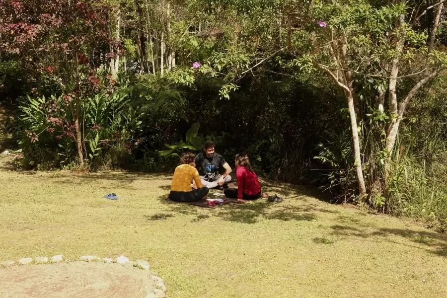 Foto de 3 pessoas sentadas em círculo, ao ar livre, na sombra de uma árvore