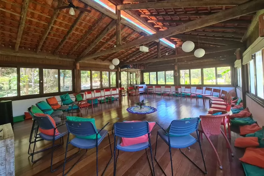 Foto grande sala clara, com piso de madeira, cheia de janelas e com cadeiras azuis, com almofadas, dispostas em círculo