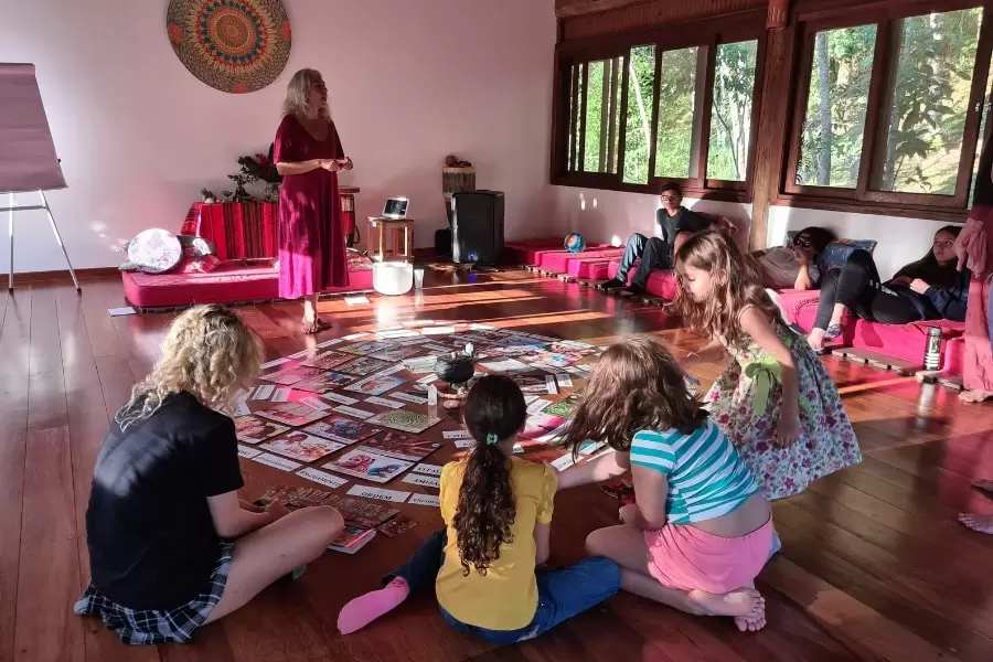 Foto de crianças sentadas em círculo, no chão de uma sala com piso de madeira, ouvindo explicações de uma mulher de pé no fundo da sala