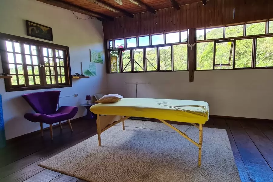 Foto de uma sala com janelas altas e com uma maca amarela posicionada ao centro , sobre um tapete