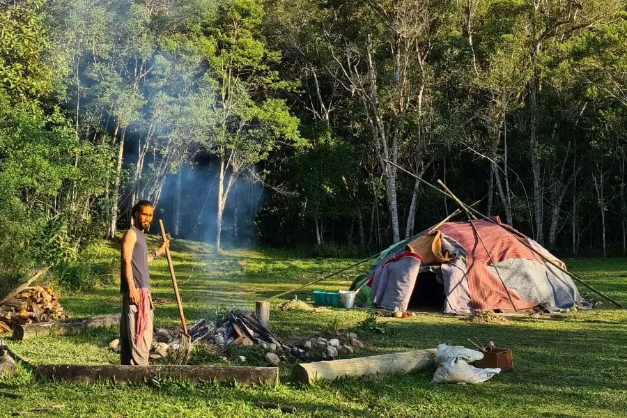 Foto de homem mexendo nas brasas de uma fogueira em frente a uma espécie de cabana