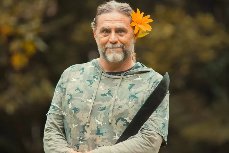 Foto de senhor com cabelo preso, uma flor laranja atrás da orelha e um facão na mão