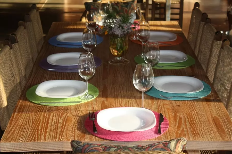 Foto de uma mesa posta, com susplats colotidos, pratos quadrados brancos e taças transparentes, numa grande mesa de madeira