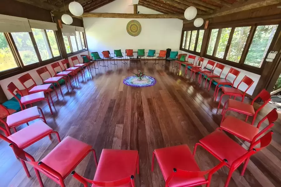 Foto grande sala clara, com piso de madeira, cheia de janelas e com cadeiras vermelhas dispostas em círculo