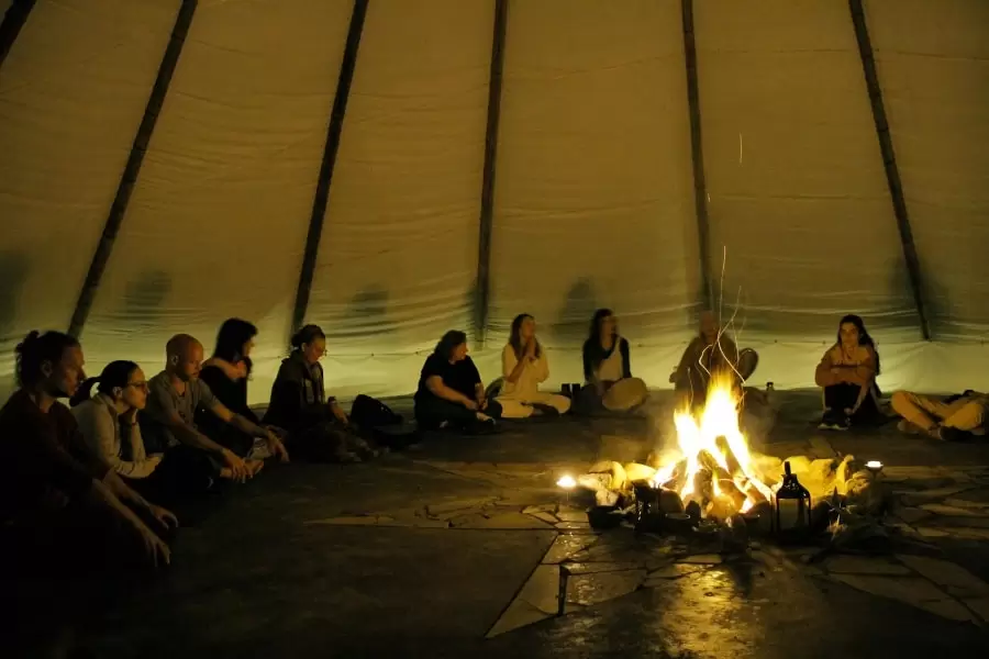 Foto de uma grande tenda, com uma fogueira no centro e pessoas sentadas no chão, em círculo, em volta dela.