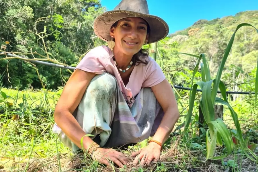 Foto de uma mulher agachada, de chapéu, com as mãos na terra