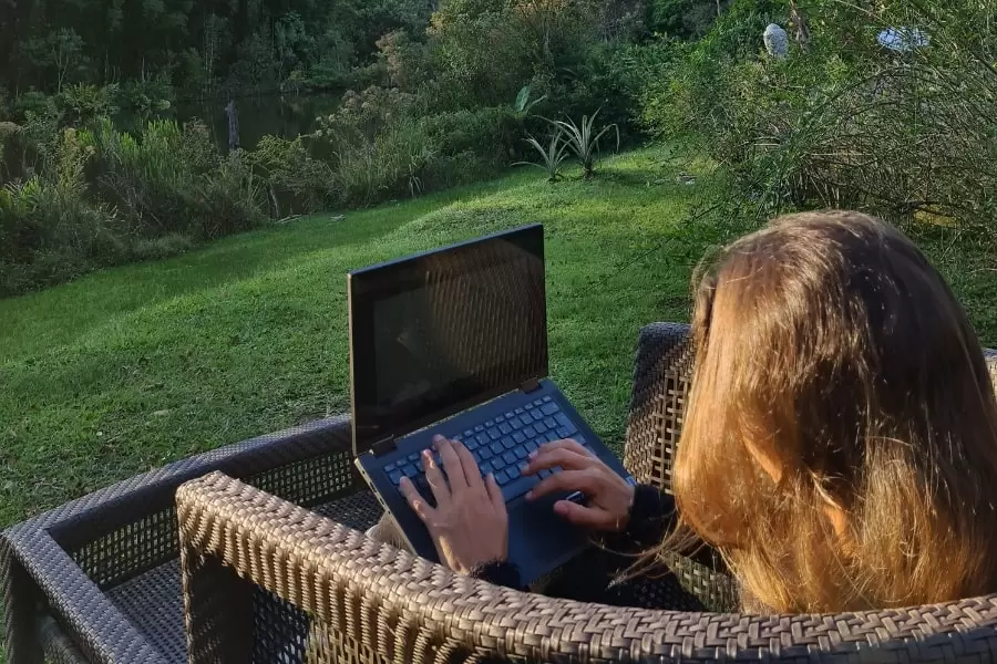 Foto de uma mulher, sentada em uma cadeira de vime, trabalhando em seu notebook