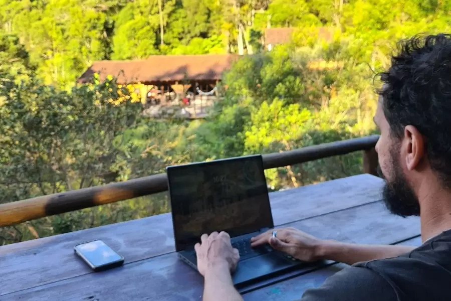 Foto de um homem, sentado em uma mesa de madeira, numa varanda com uma paisagem de mata, trabalhando em seu notebook