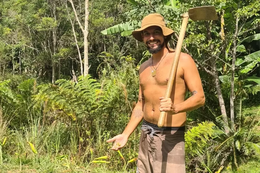 Foto de um homem de chapéu, no sol, com um enxada no ombro e a mata ao fundo